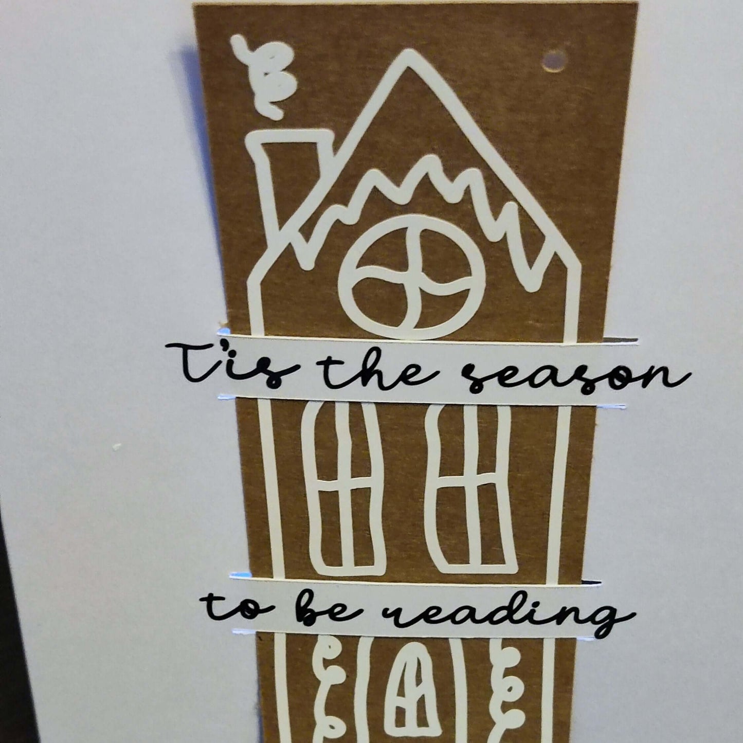 Close-up of a handmade Christmas card featuring a tall gingerbread-style house in white on a brown background. Across the house, black script text reads: '’Tis the season to be reading'.