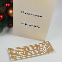 Handmade Christmas card displayed upright with black script text reading '’Tis the season to be reading'. A matching brown gingerbread-style house bookmark with white detailing lies in front of it. A festive pine branch with red and gold baubles is in the background.