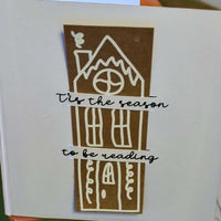 Handmade Christmas card featuring a tall gingerbread-style house in white on a brown background. Across the house, two banners read: '’Tis the season to be reading' in black cursive font.