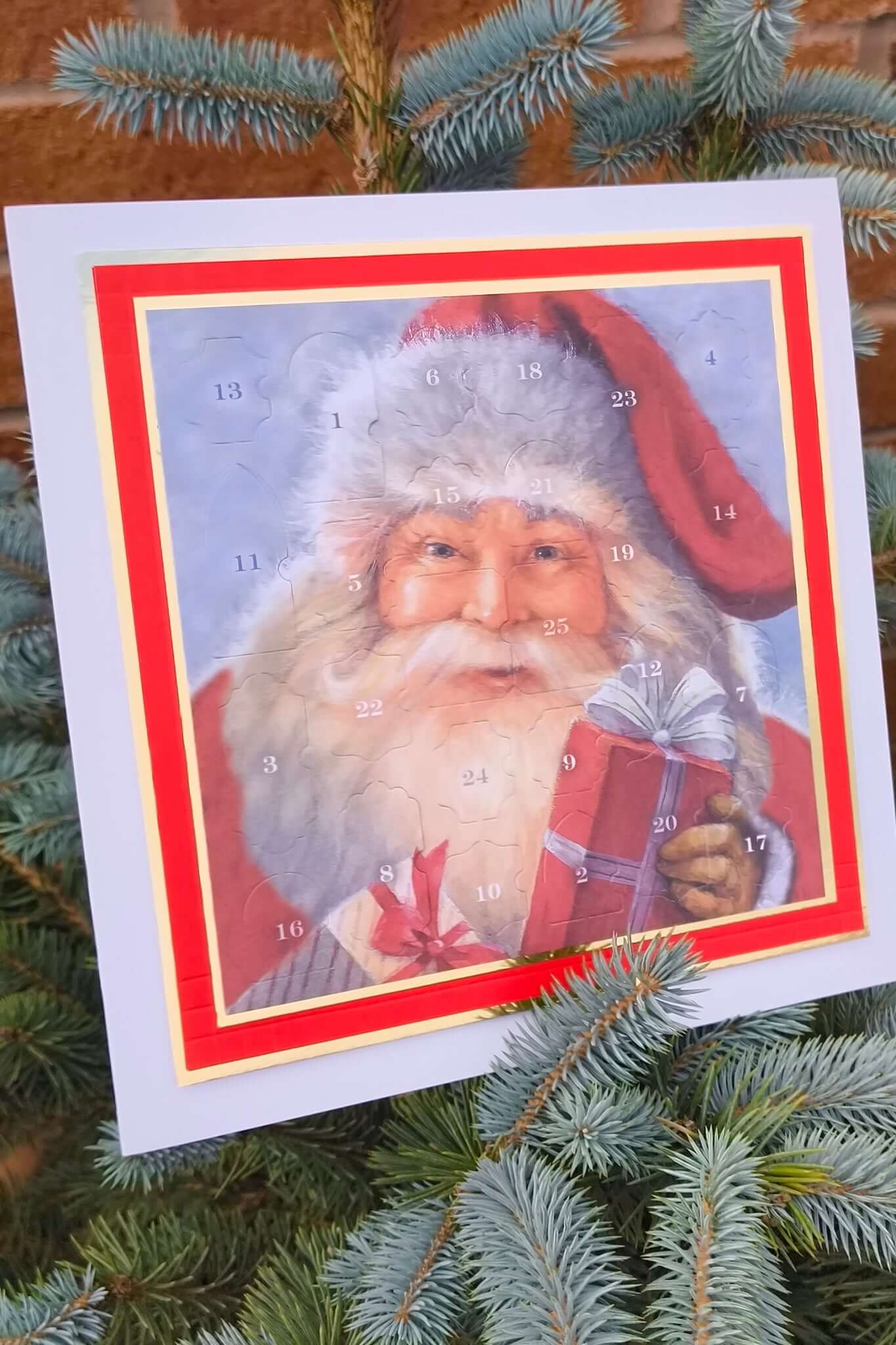 Close-up of a traditional Father Christmas Advent Calendar with numbered doors, featuring a vintage-style Santa holding a red present, displayed on a blue spruce tree outdoors.