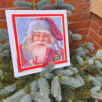 Traditional Father Christmas Advent Calendar with numbered doors, featuring a vintage Santa holding a red gift, displayed on a small outdoor Christmas tree in a terracotta pot against a brick wall.