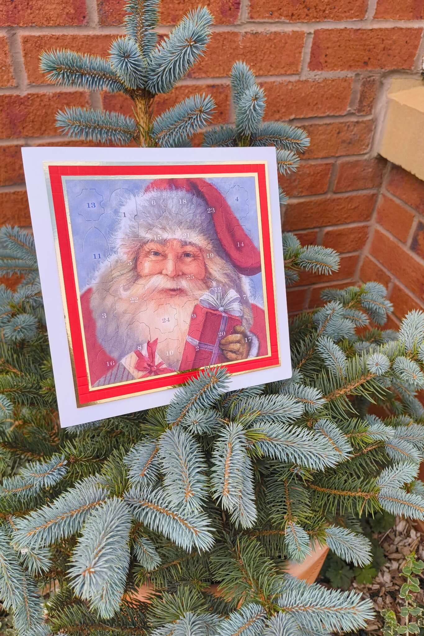 Traditional Father Christmas Advent Calendar with numbered doors, featuring a vintage Santa holding a red gift, displayed on a small outdoor Christmas tree in a terracotta pot against a brick wall.