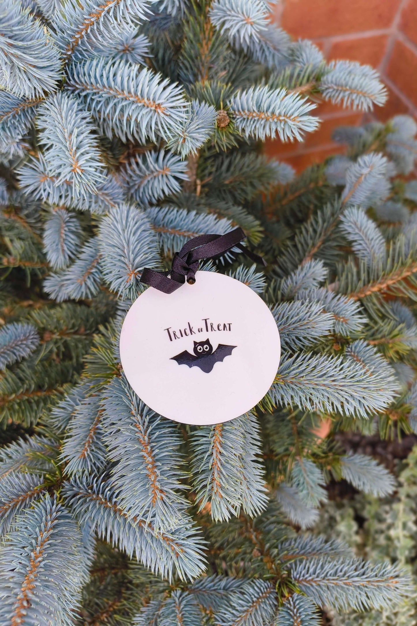Trick or Treat Halloween bauble with black bat print, hanging on outdoor evergreen branch.