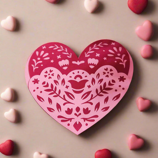 Layered heart papercut in pink and red with folk floral design, top-down flat-lay surrounded by candy hearts on neutral background, handmade Valentine’s Day card

