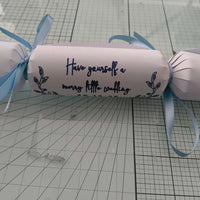 White cracker with navy foil wedding lettering “Have yourself a merry little wedding,” tied with pale blue ribbon on craft background.