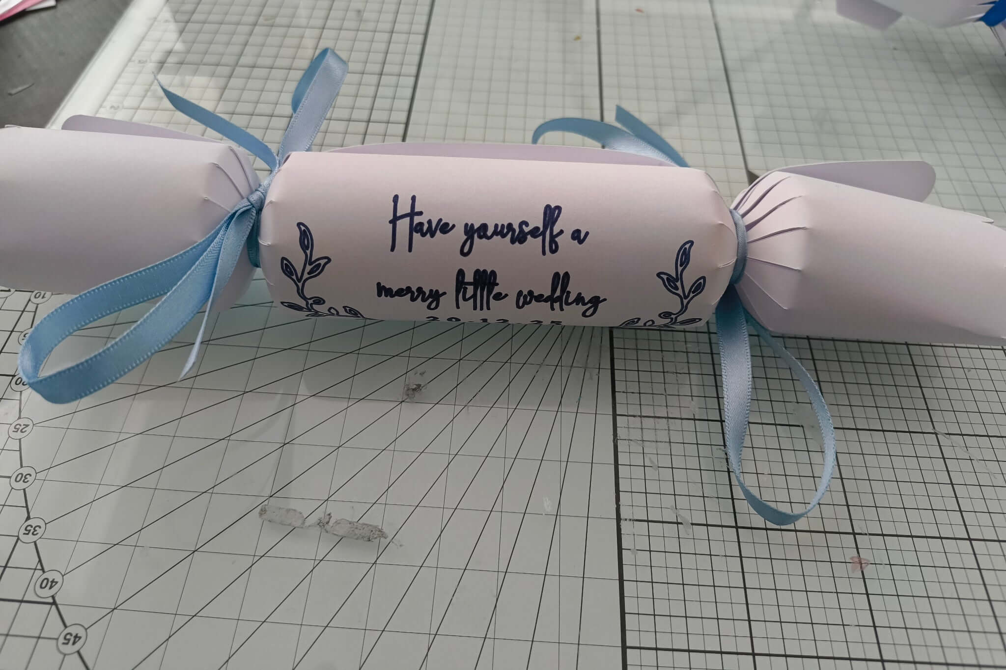 White cracker with navy foil wedding lettering “Have yourself a merry little wedding,” tied with pale blue ribbon on craft background.