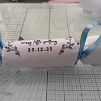 White cracker with navy foil wedding lettering and date, finished with pale blue ribbons, styled flat on craft mat background.