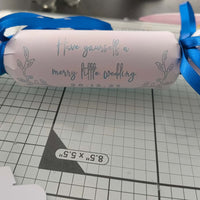 White cracker with silver foil message “Have yourself a merry little wedding” and date, tied with royal blue satin ribbon, styled on a craft mat.