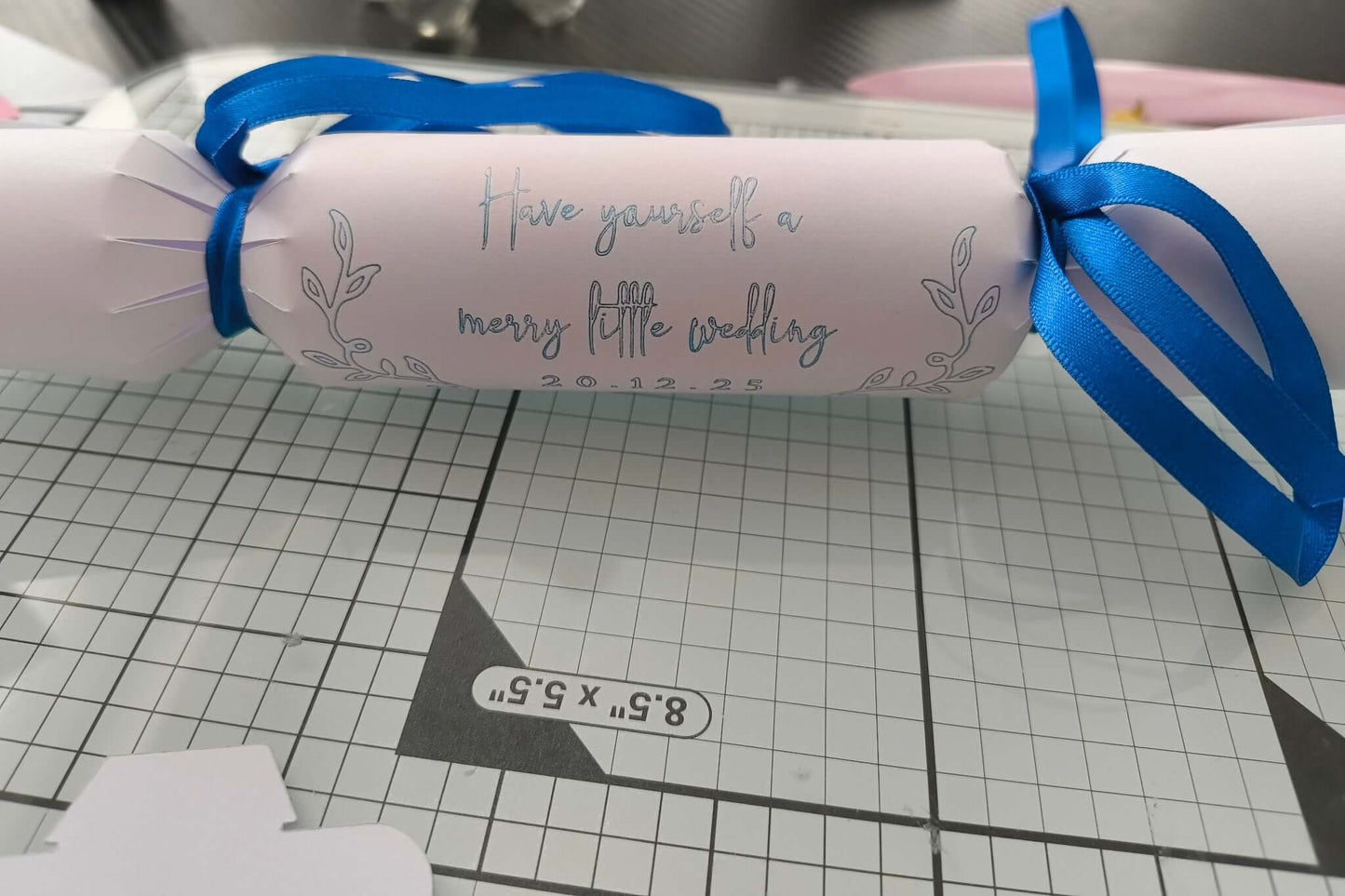 White cracker with silver foil message “Have yourself a merry little wedding” and date, tied with royal blue satin ribbon, styled on a craft mat.