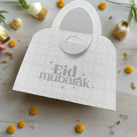 White Eid Mubarak handbag-shaped money holder with delicate floral detail, surrounded by yellow flowers, perfect Eid gift idea.