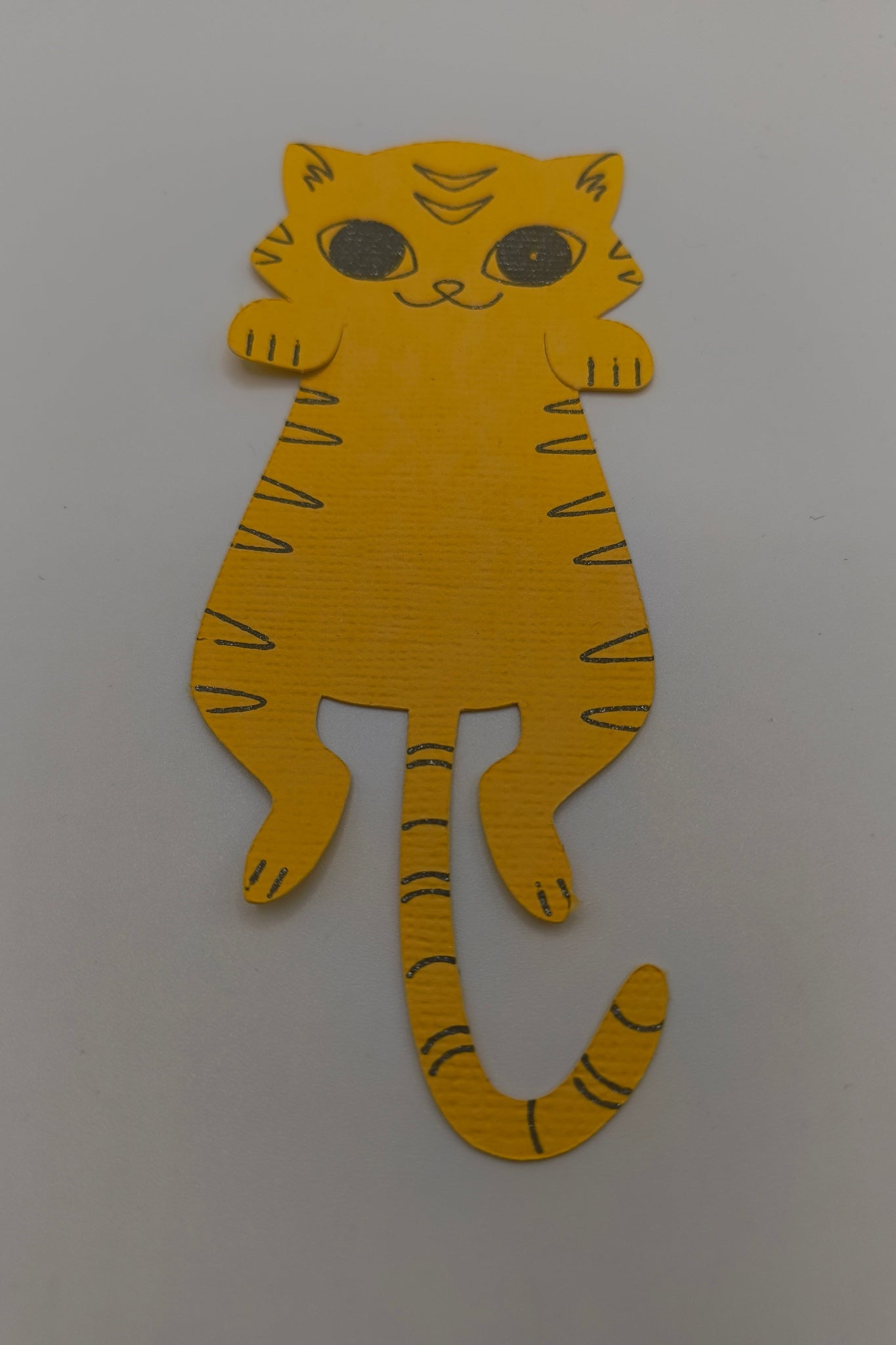 Handmade yellow paper cat cut-out with drawn stripes, big eyes, and a long tail on a white background.